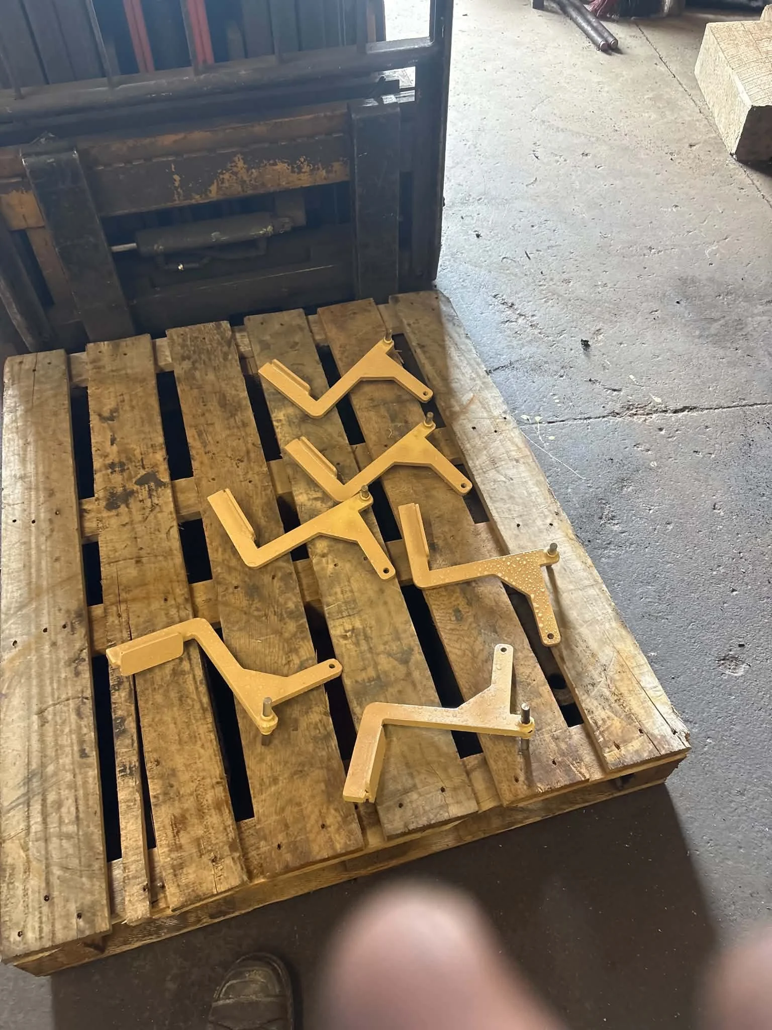 Several beige metal L-shaped brackets or hangers placed on a wooden pallet with a tool shadow visible at the bottom of the image.