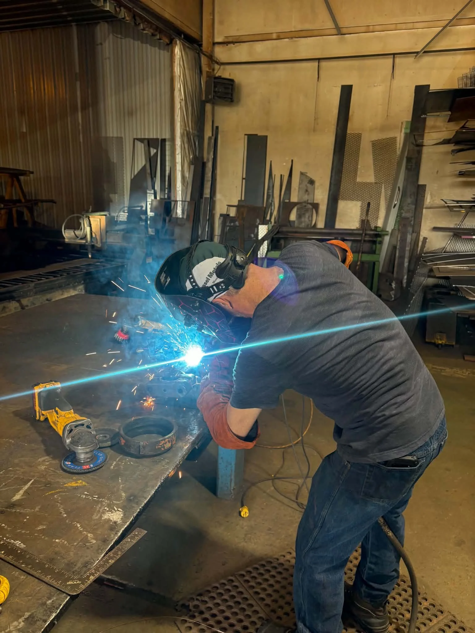 A person welding metal parts in a workshop, wearing a welding helmet, with sparks and bright blue light from the welding process, various tools and metal pieces on a workbench.