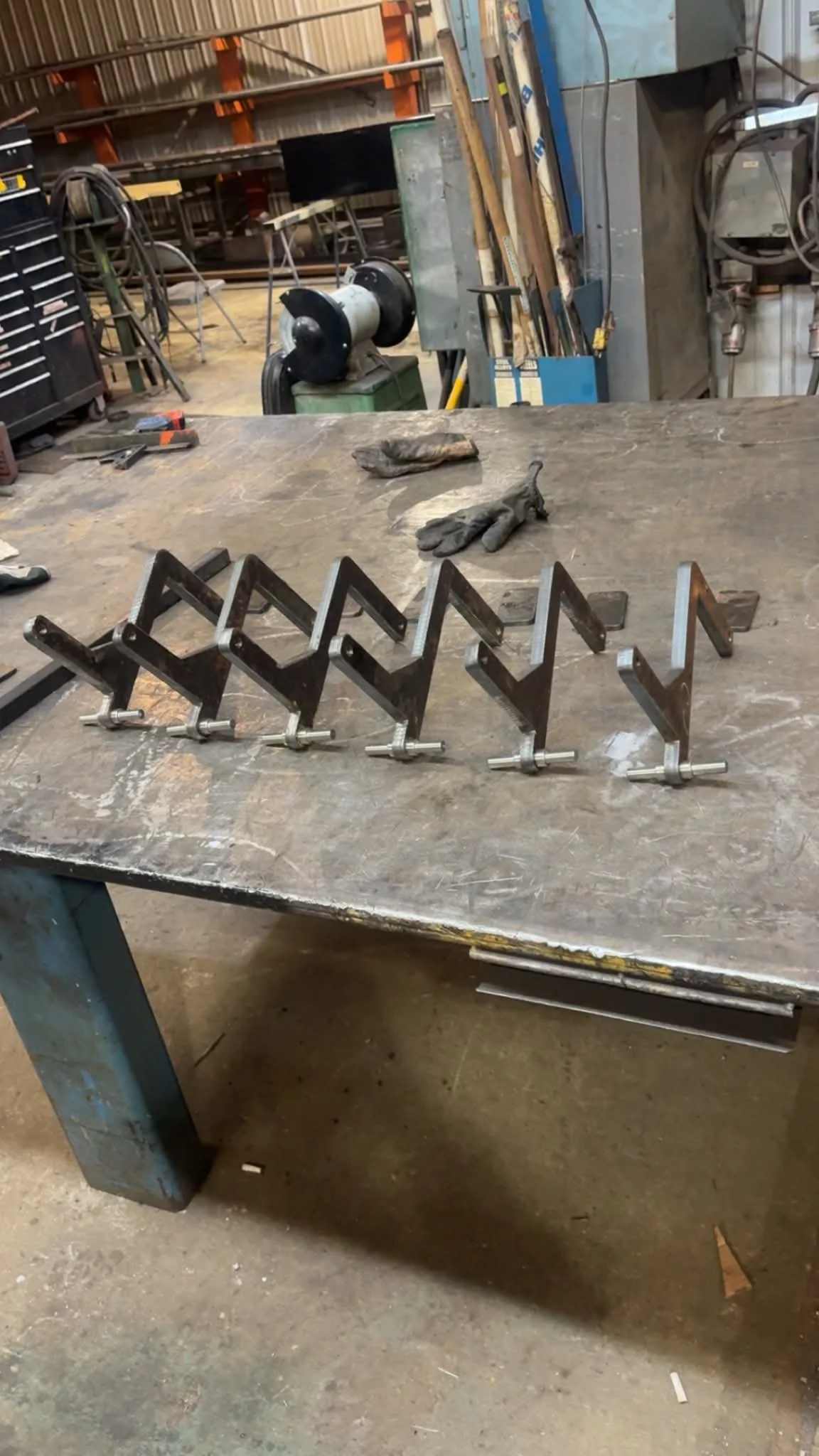 A metal zigzag clamp on a worktable in a workshop, with gloves and tools in the background.
