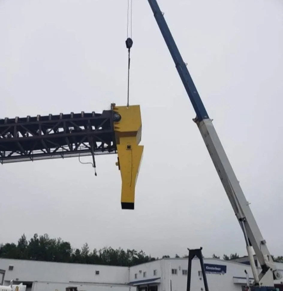 A large crane lifting a yellow piece of equipment at a construction site with a gray overcast sky.