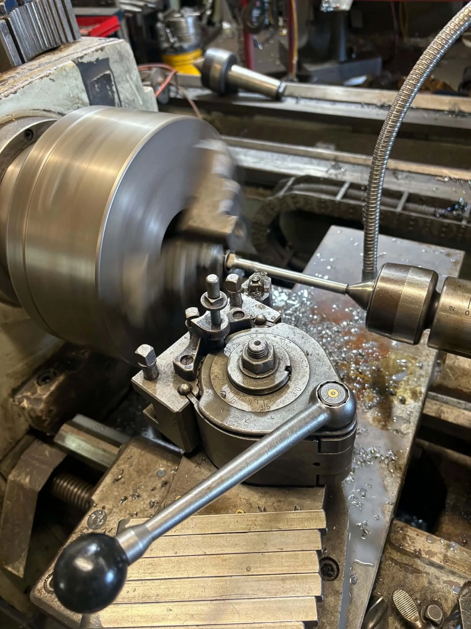 A metal lathe machine in operation shaping a cylindrical metal workpiece with chips and coolant around. Various machine parts and tools are visible in the background.