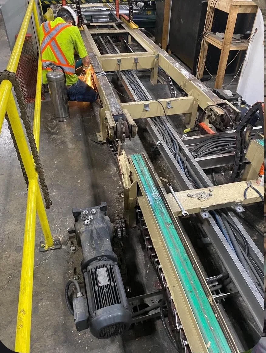 A worker in a yellow safety vest and helmet welding on a large industrial machine frame in a manufacturing facility.