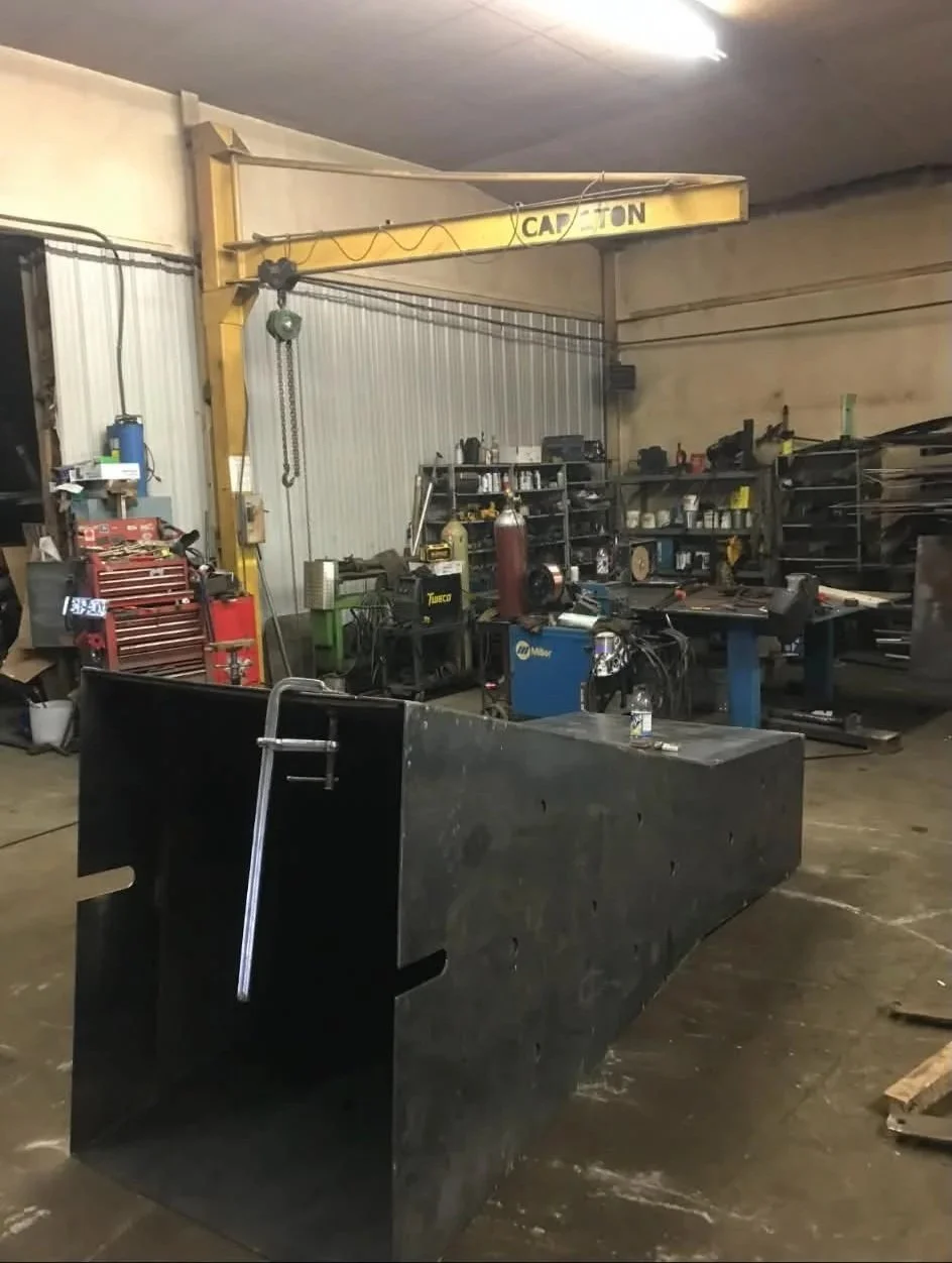 A workshop with a black metal workbench in the foreground, shelves with tools and equipment in the background, and a yellow overhead crane labeled 'CAUTION' hanging from the ceiling.
