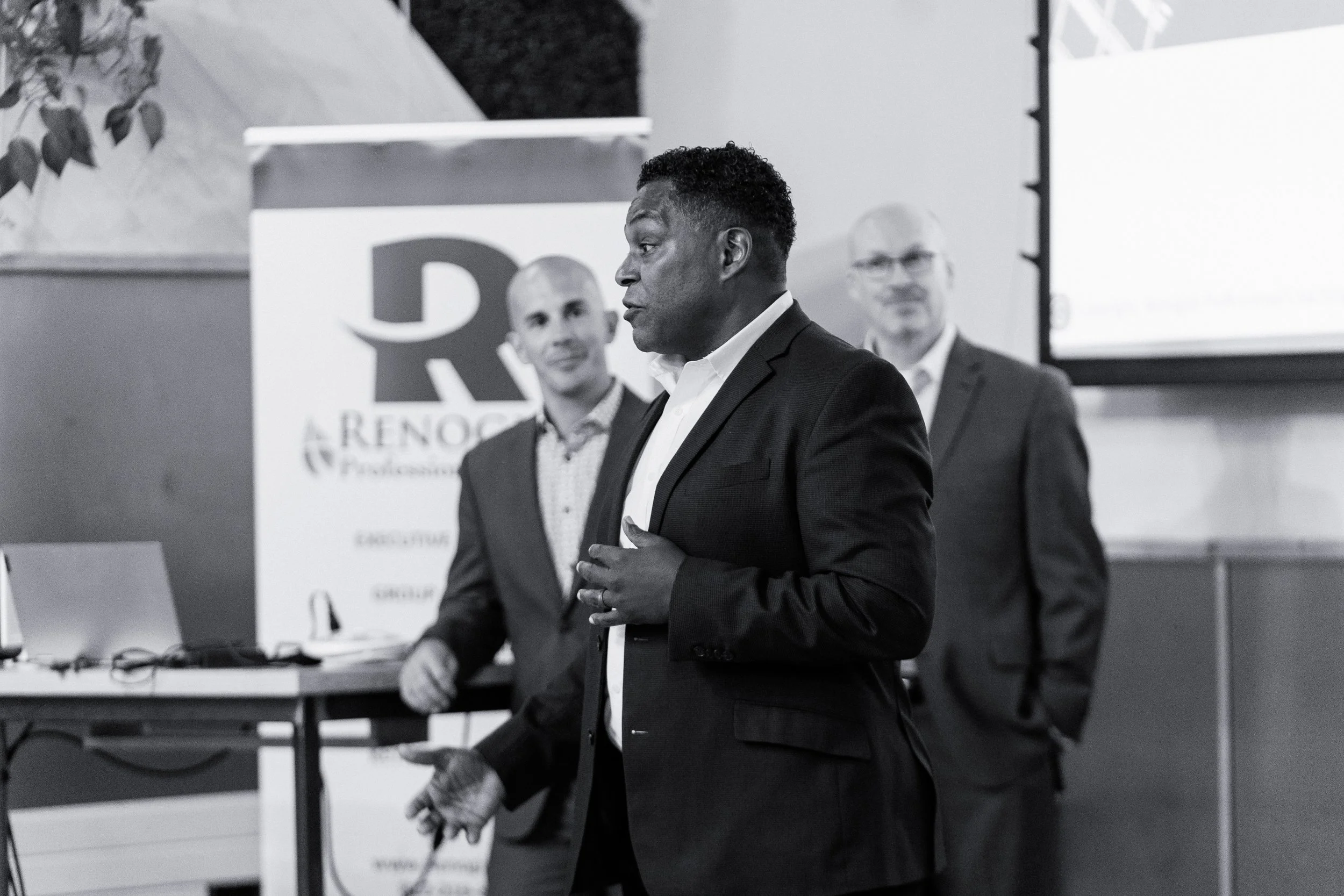 Man in a suit giving a presentation, with two men in suits, one with glasses, standing behind him in a conference or event setting.