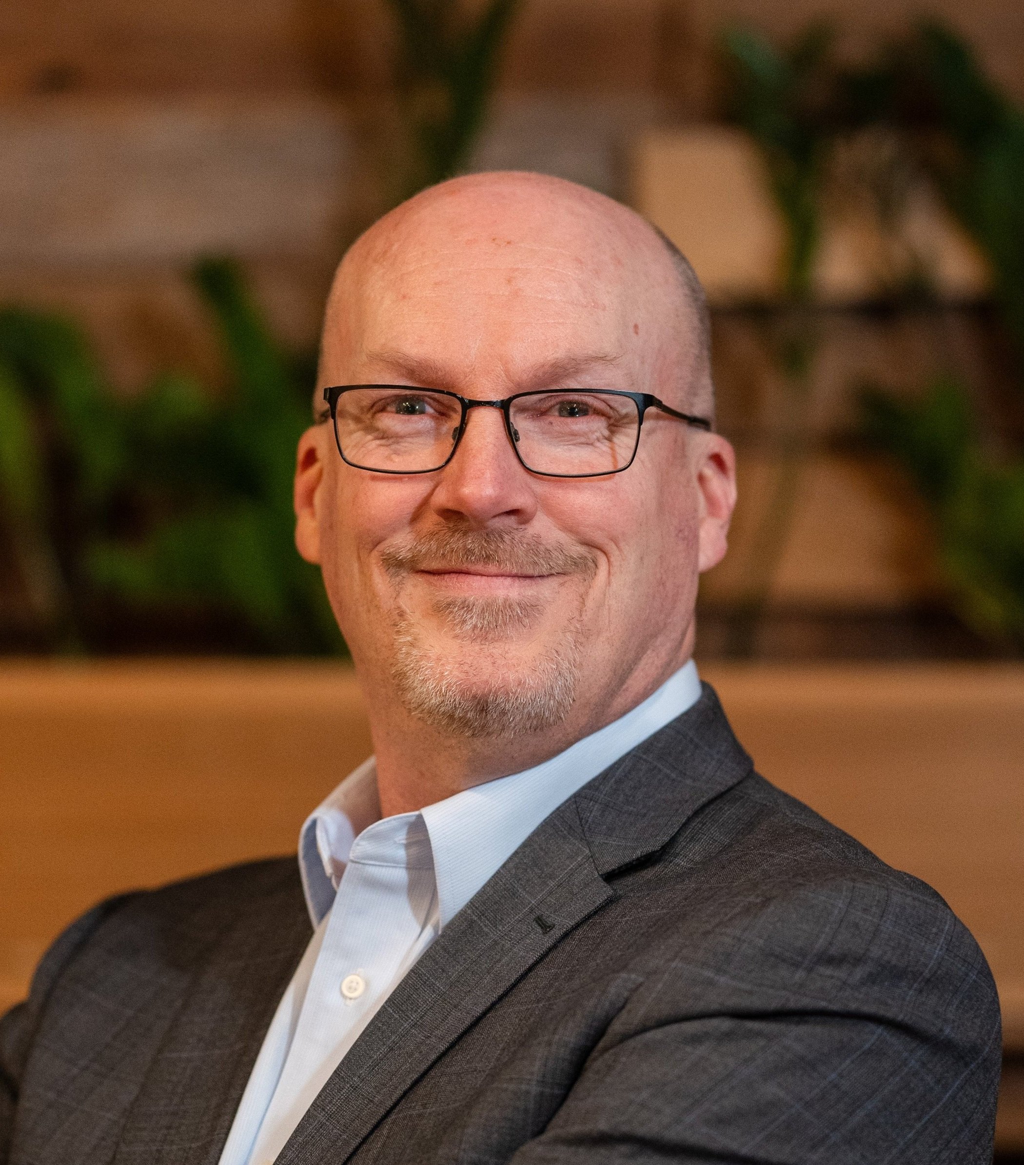 A middle-aged man with glasses and a bald head smiling, wearing a dark checkered suit jacket and a light blue dress shirt, in front of a blurred background with green foliage.