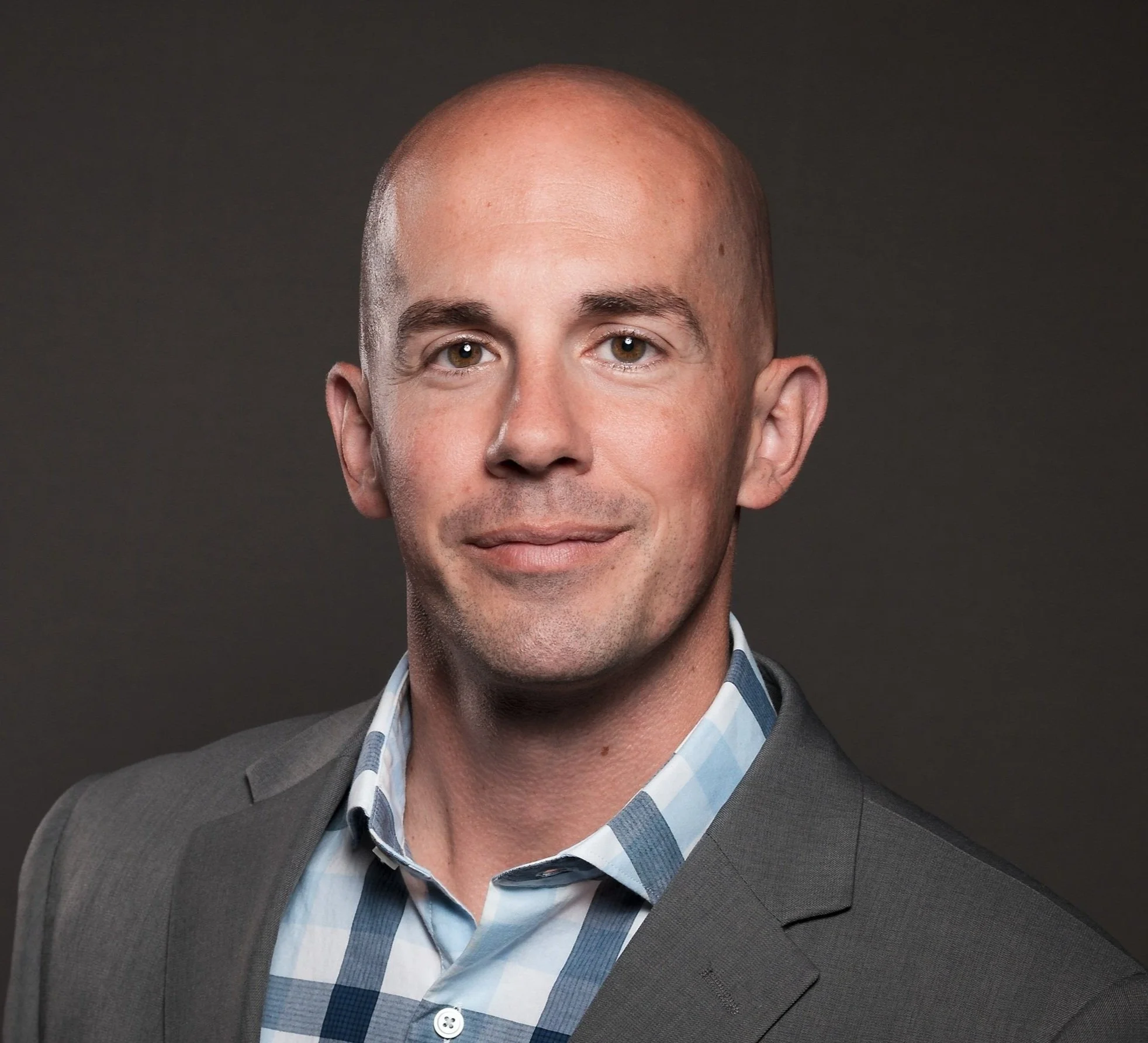 Close-up of a smiling man with a bald head, dressed in a gray blazer and a blue checkered shirt, against a dark background.