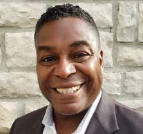 Portrait of a smiling man with short hair, wearing a dark blazer and a collared shirt, standing against a stone wall background.