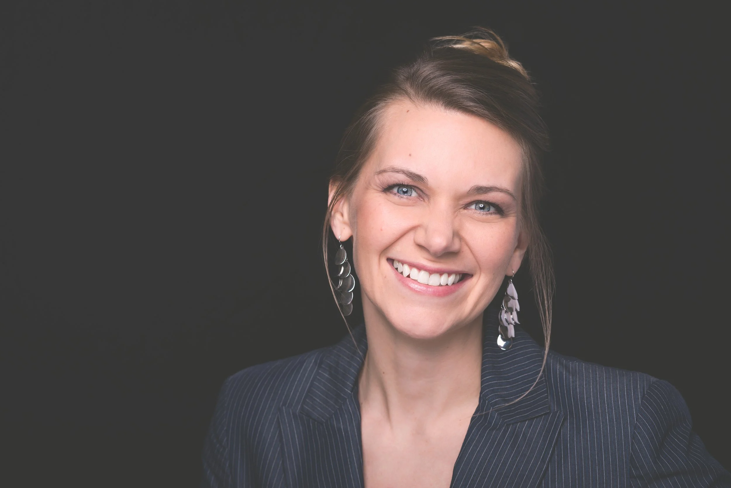 Close-up of a woman with blue eyes, smiling, wearing earrings and a dark pinstripe blazer against a black background.