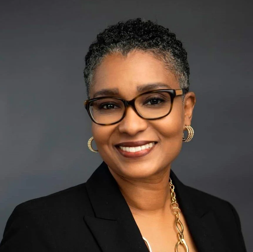 A smiling woman with short curly gray hair, wearing black glasses, hoop earrings, a black blazer, and a gold chain necklace against a dark gray background.
