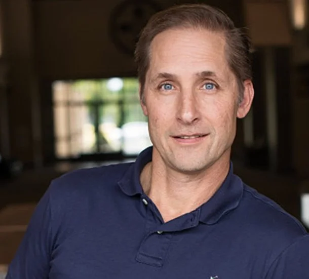 Portrait of a middle-aged man with light skin, blue eyes, and brown hair wearing a navy blue polo shirt, in an indoor setting with windows in the background.
