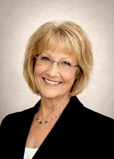 Smiling middle-aged woman with blonde hair, glasses, wearing a black blazer and necklace.