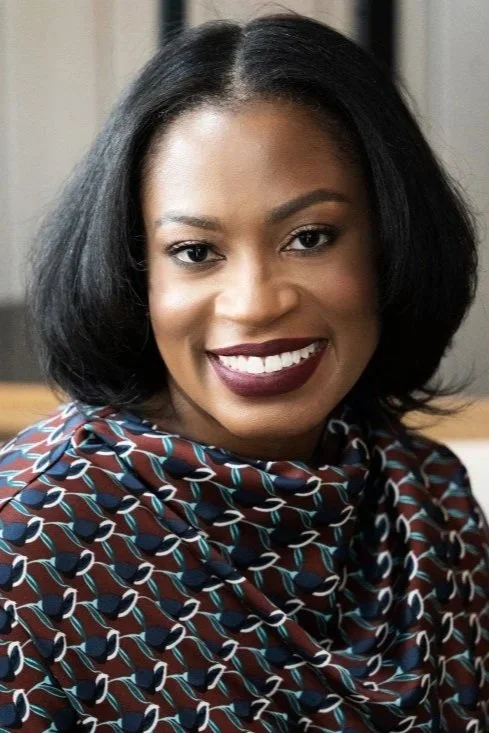 A smiling woman with shoulder-length black hair, wearing a patterned blouse with blue, white, and brown colors, indoor setting with a window in the background.