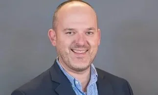 Headshot of a smiling man with short hair, wearing a suit jacket and a blue collared shirt, against a plain background.