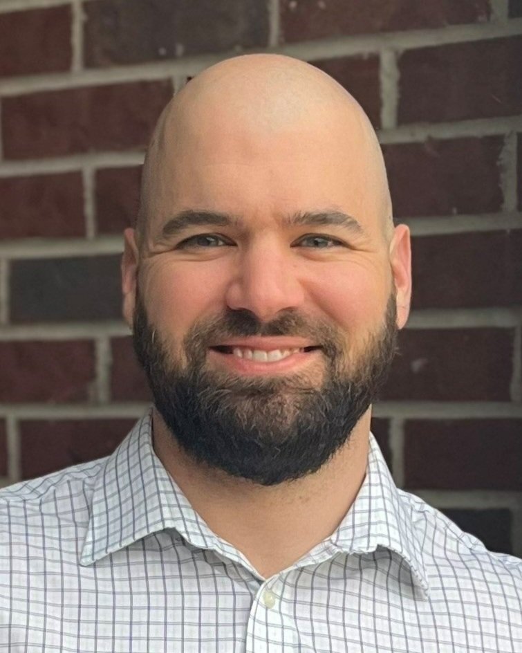 A man with a beard, smiling, wearing a checkered shirt, standing in front of a brick wall.