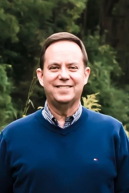 A smiling man in a blue sweater and checkered shirt standing outdoors with a background of trees.