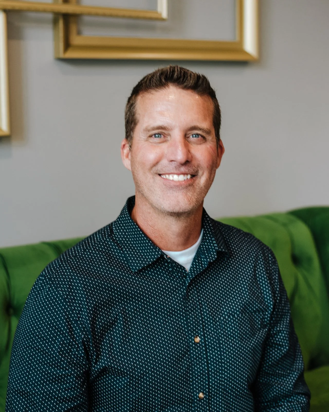 A smiling man with short brown hair and blue eyes sitting on a green couch with a gray wall and picture frames in the background.