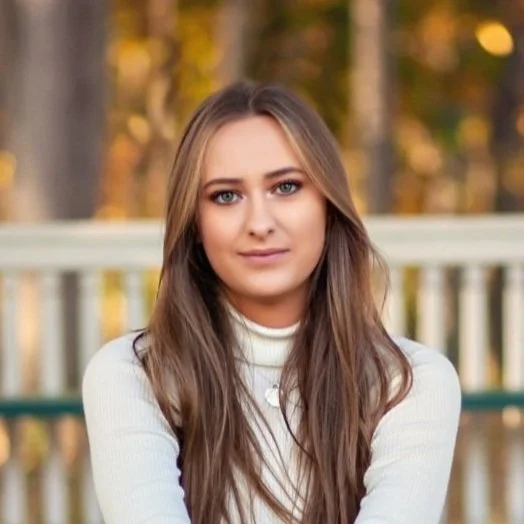 A young woman with long brown hair and blue eyes sitting outdoors on a wooden bridge in a park with autumn leaves in the background.
