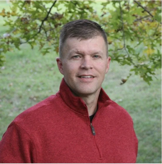 A man with short brown hair wearing a red zip-up jacket, standing outdoors under a tree with green leaves.