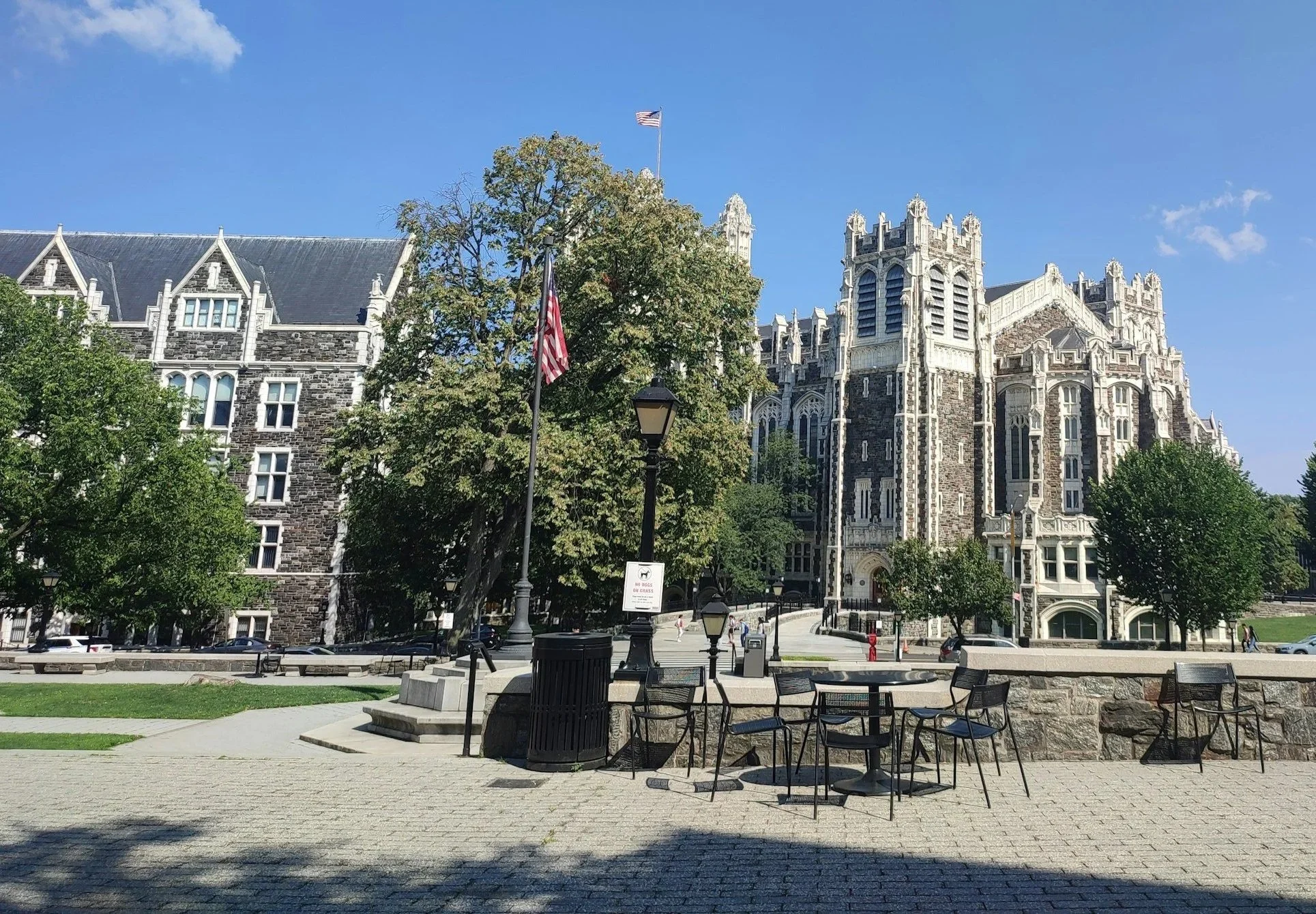 A historic stone building with gothic architecture, including multiple towers and arched windows, under a blue sky with few clouds. In front, a park with trees, benches, and black street lamps, along with an American flag flying on flagpoles.