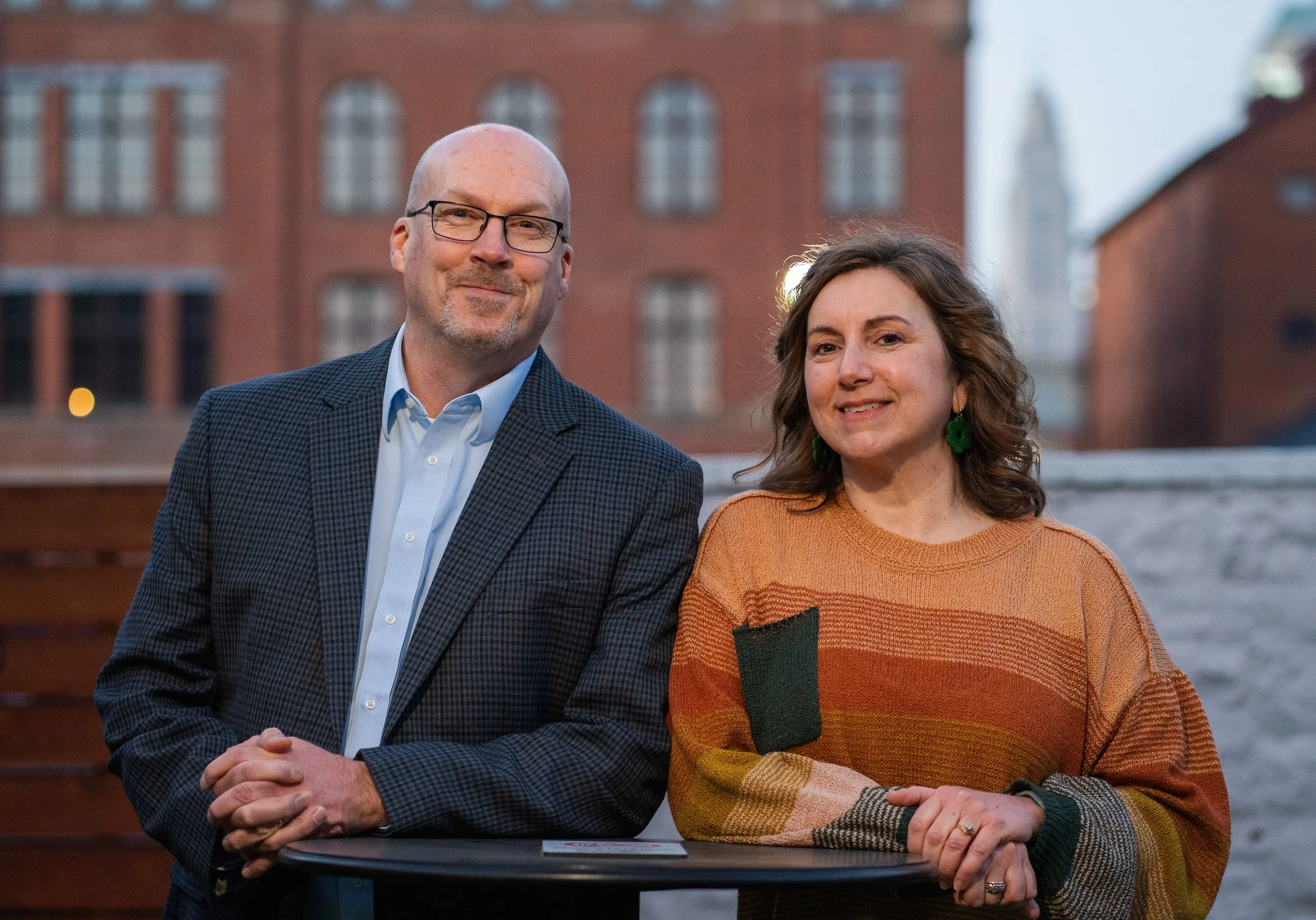 A man with glasses and a woman with curly hair standing outdoors near a body of water with brick buildings in the background.