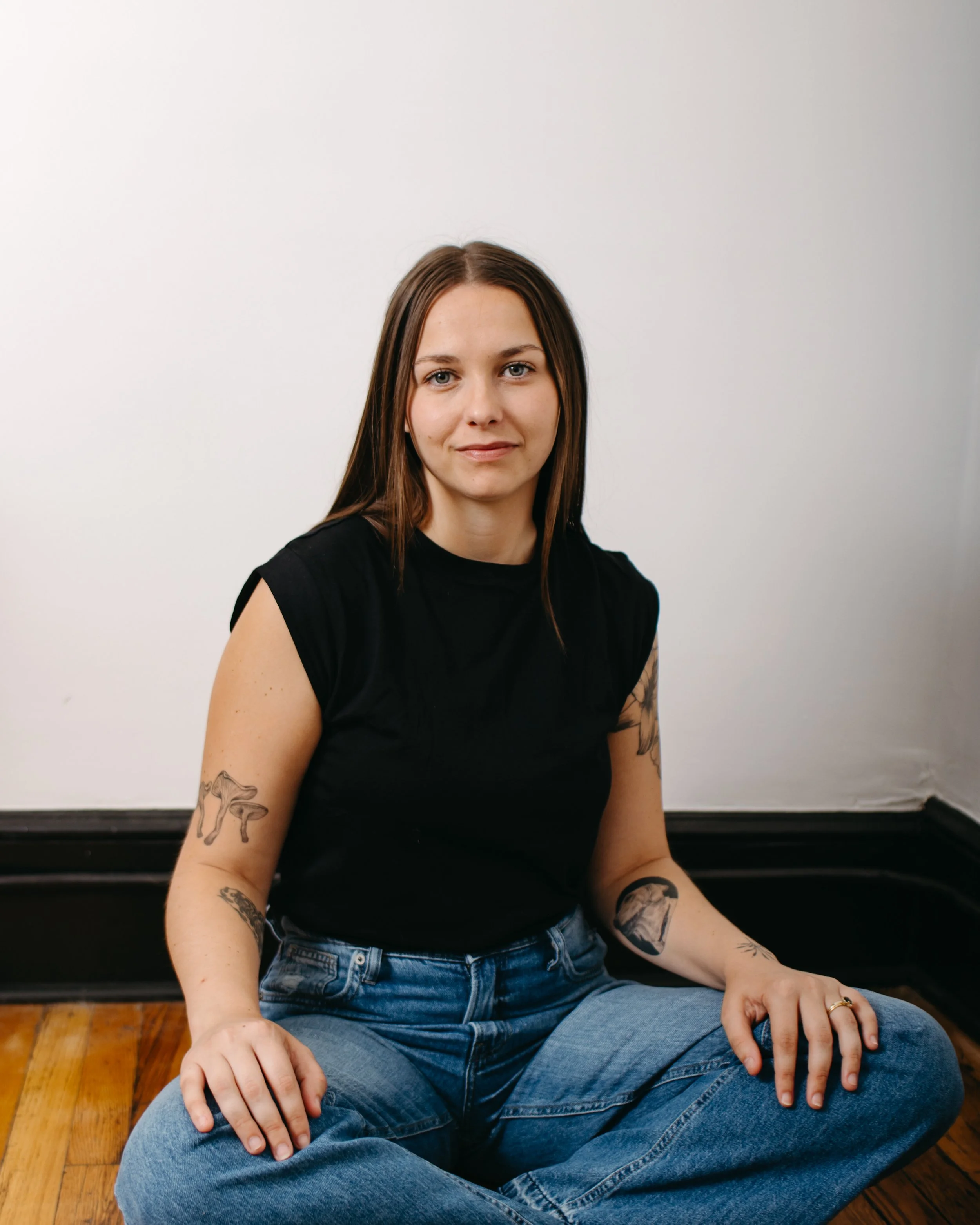 A woman with long brown hair and tattoos on her arms sits cross-legged on a wooden floor, wearing a black shirt and blue jeans, against a plain white wall.