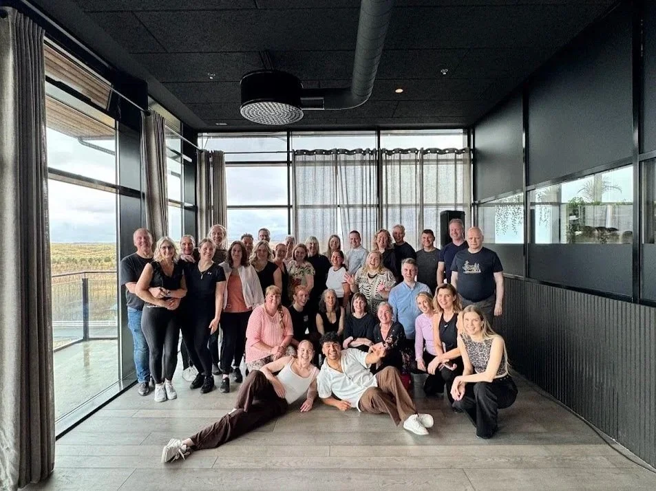 Group of people gathered inside a modern room with large windows and curtains, posing for a photo and smiling.