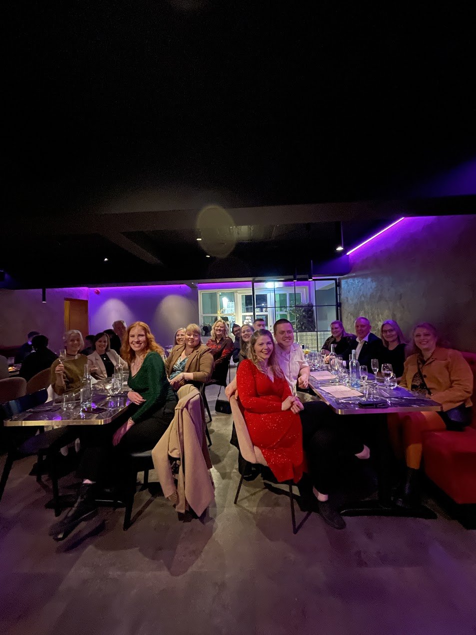 Group of people sitting at a long table in a restaurant with dim lighting and purple neon accents, smiling and enjoying their meal.