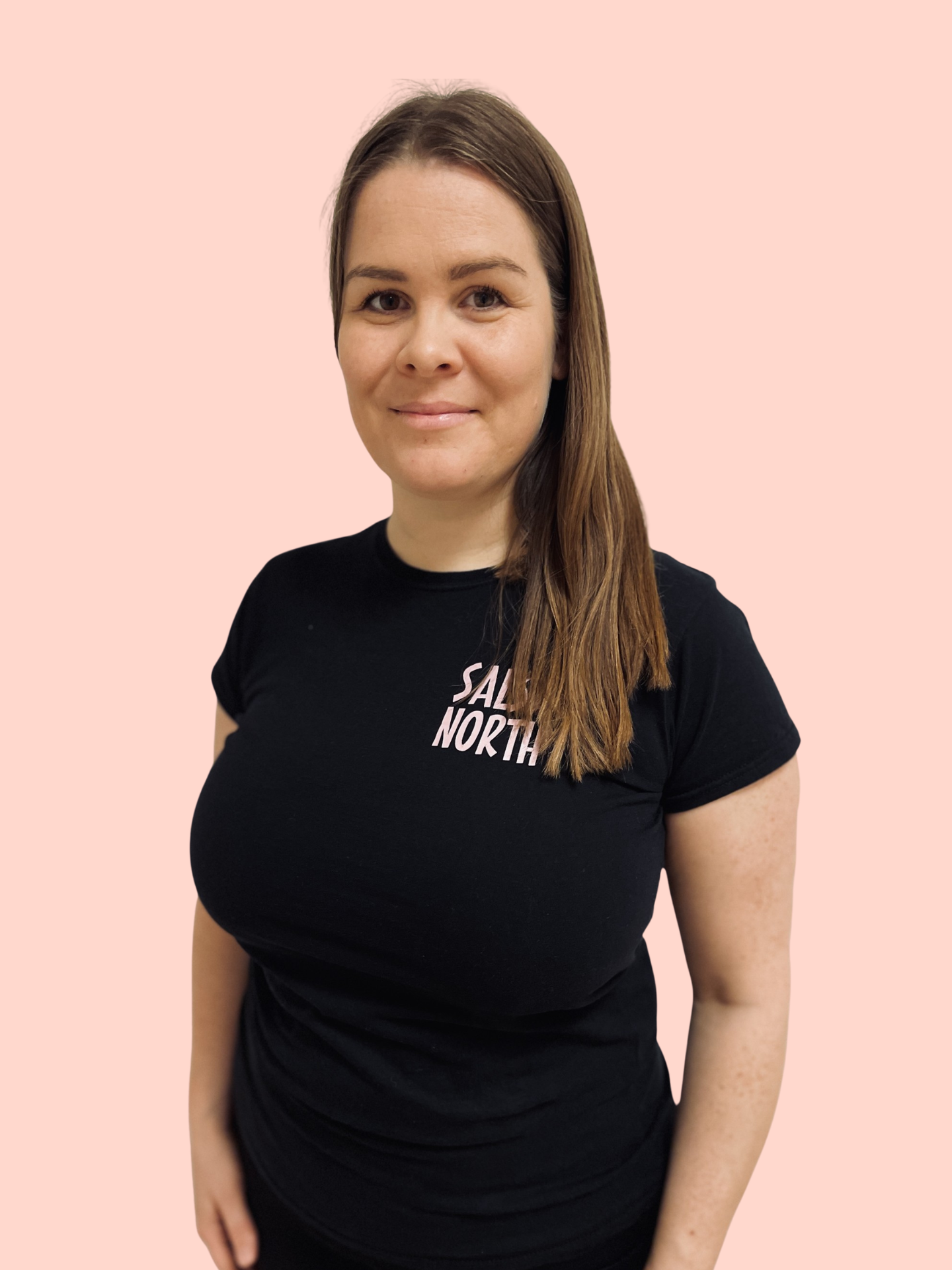 A woman with long brown hair wearing a black T-shirt standing against a light pink background.