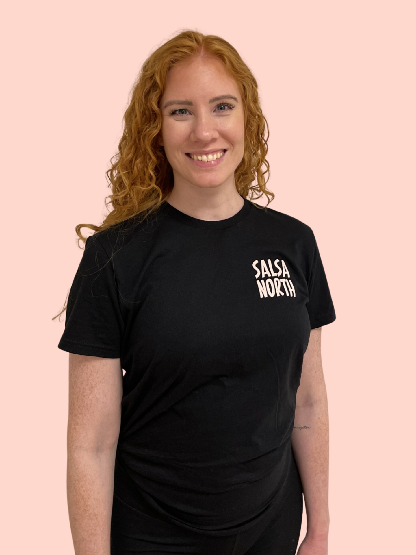 A woman with long curly red hair wearing a black T-shirt with 'SALSA NORTH' written on it, smiling against a light pink background.
