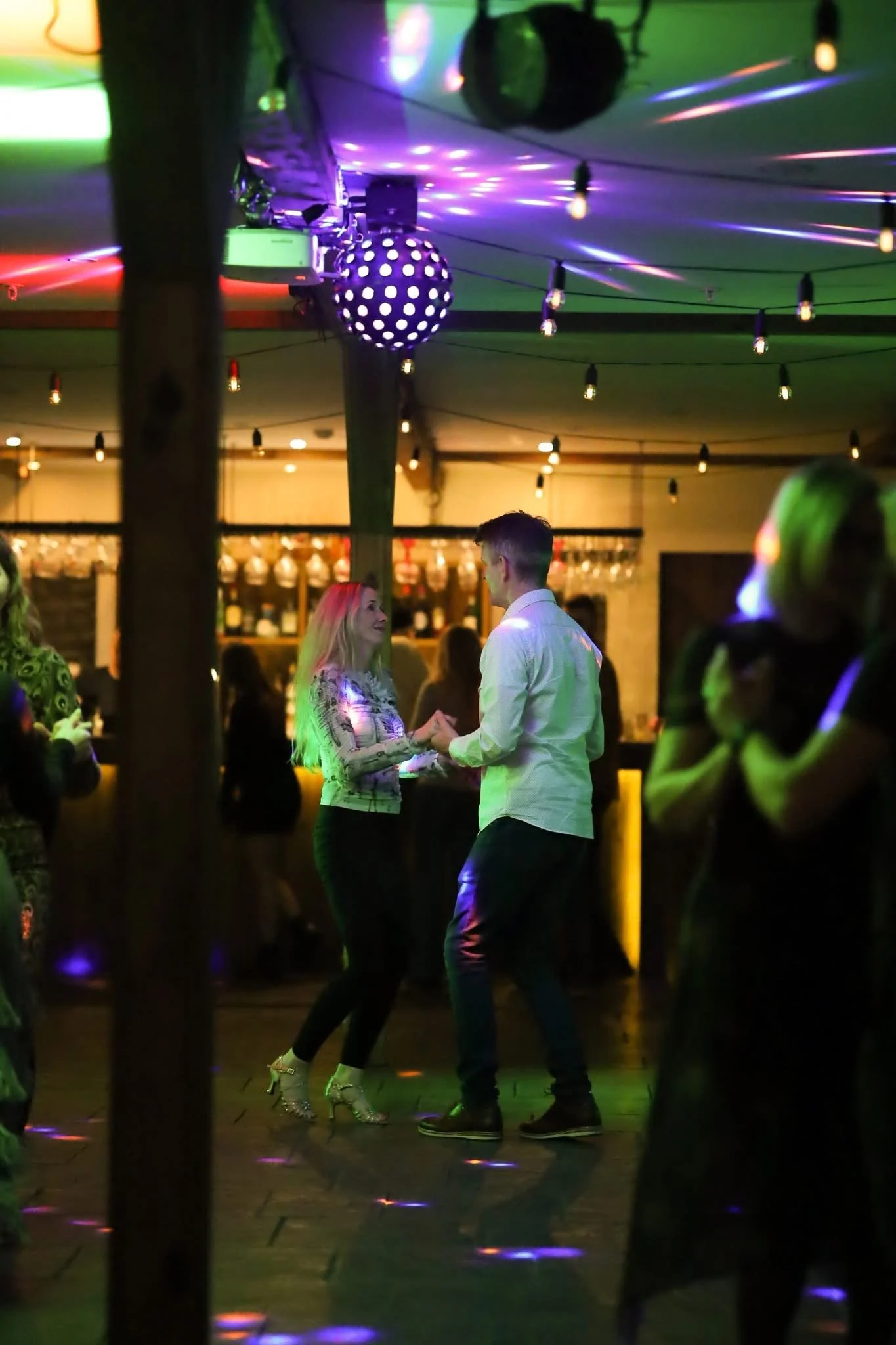 A couple dancing under colorful disco lights in a lively indoor setting, with string lights hanging from the ceiling and people in the background.