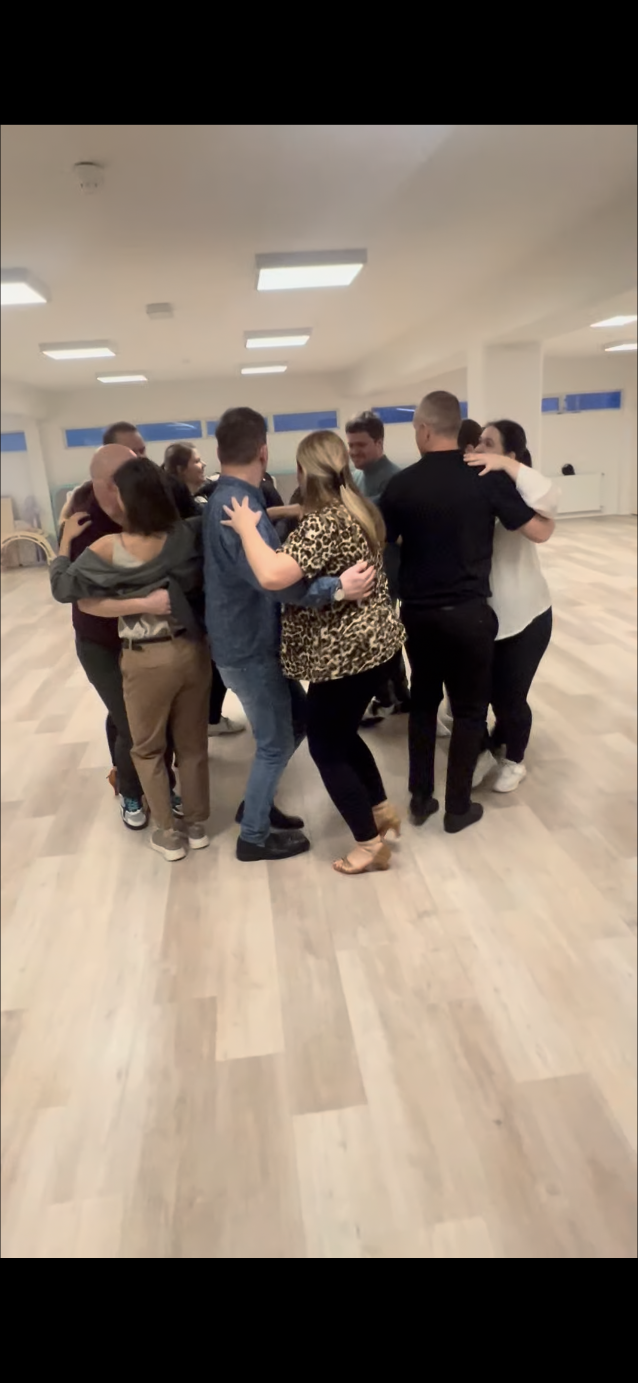 A group of people in casual and semi-formal clothing are hugging and dancing in a spacious, well-lit room with wooden floors.