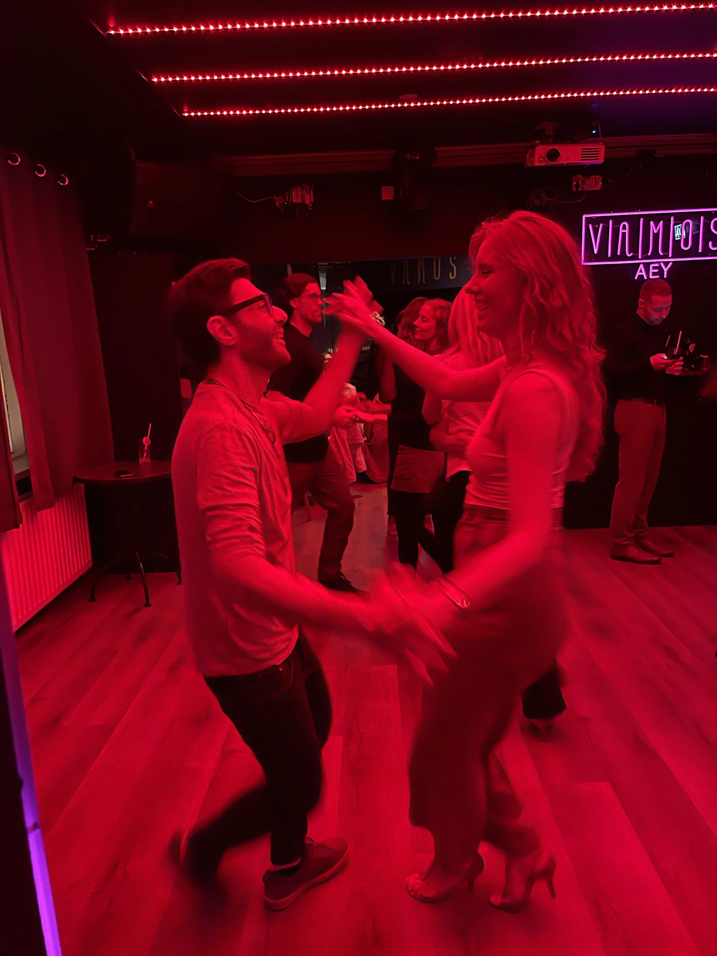 People dancing in a dimly lit nightclub with red lighting, neon signs, and a wooden floor.