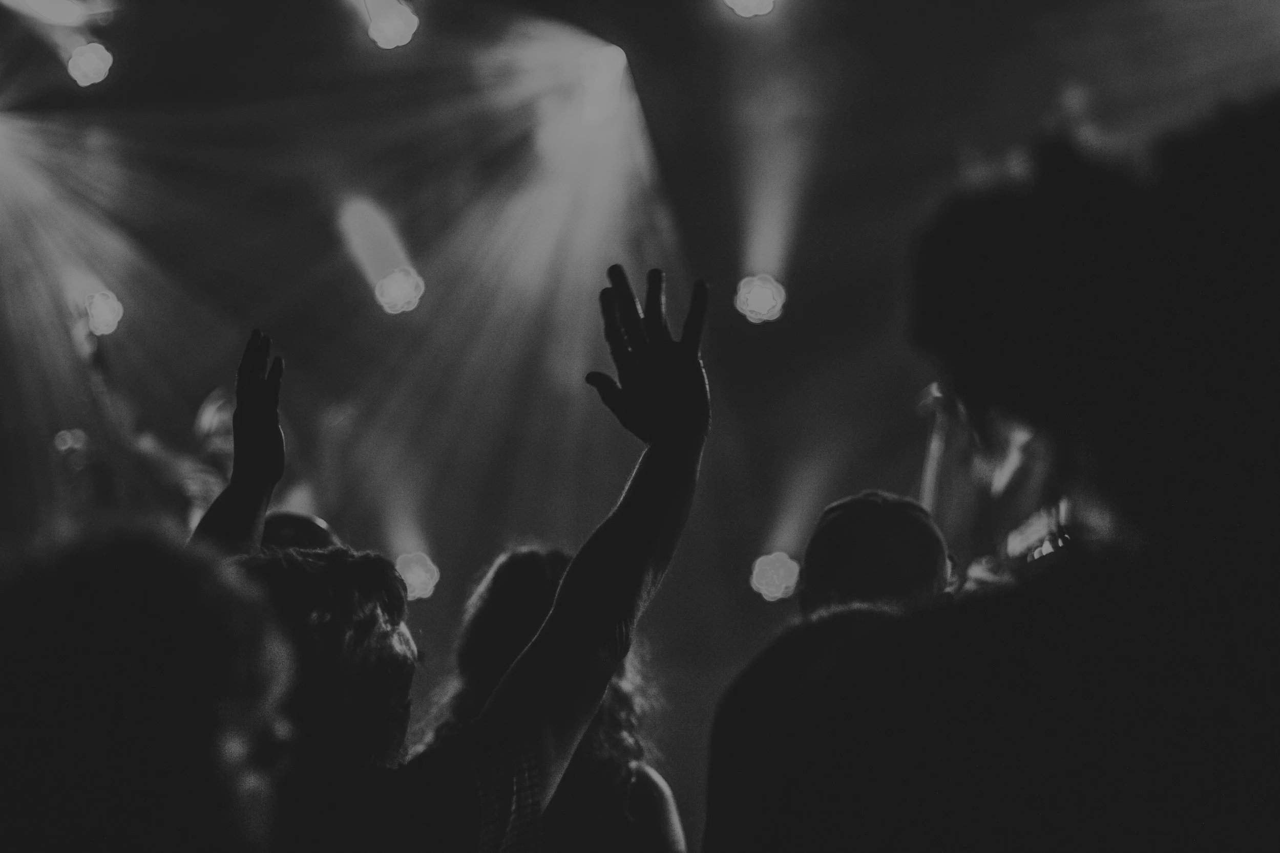 Black and white photo of a crowd at a concert, with hands raised and stage lights creating a dramatic atmosphere.