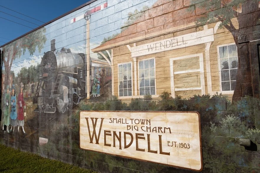 Murale of a historic train station in Wendell with a vintage train, people, and buildings, featuring a sign that reads "Small Town Big Charm Wendell Est. 1903."