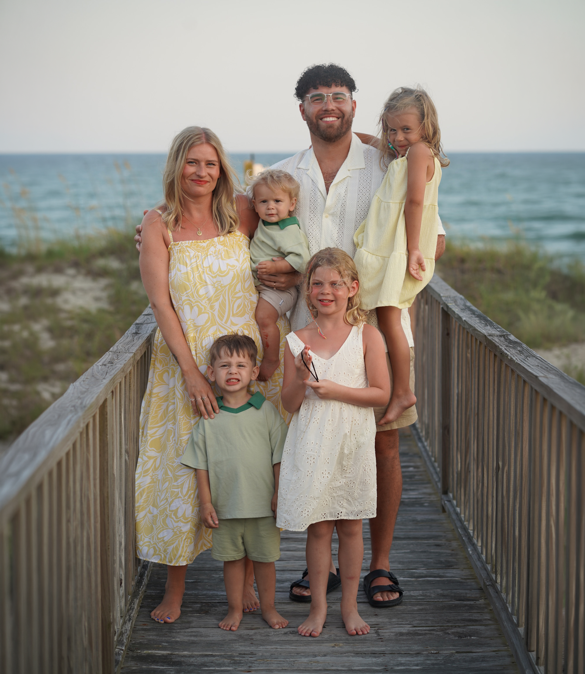 A group of seven people, including three children and four adults, standing on a wooden boardwalk or bridge by the water, with a beach and water in the background, during daytime.