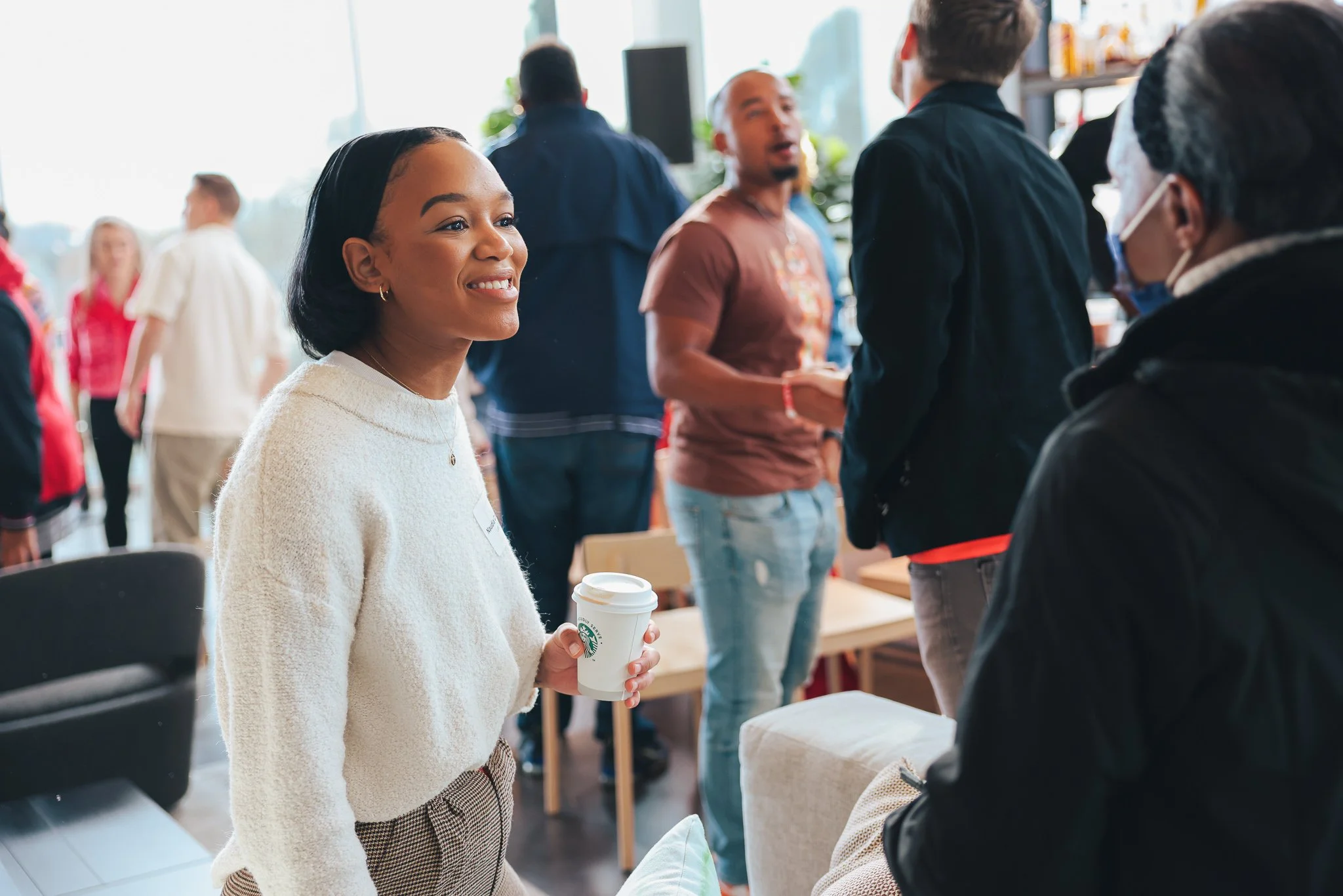 A woman with a warm smile holding a Starbucks coffee cup engaging in conversation with a man wearing glasses and a face mask at a social gathering.