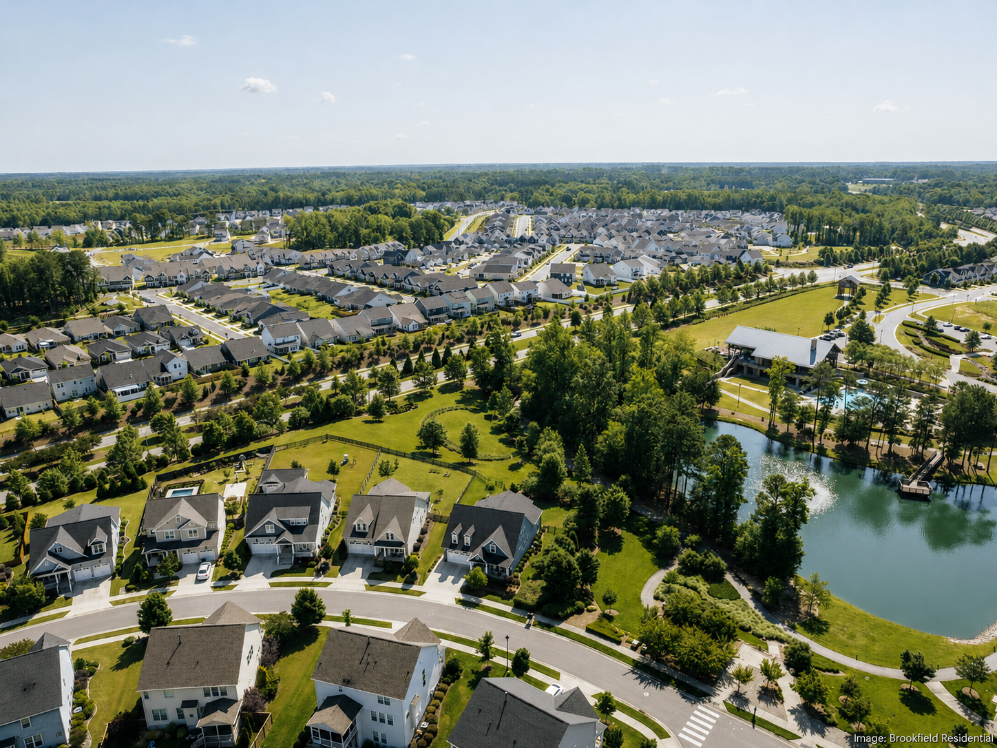Aerial view of a suburban neighborhood with houses, green lawns, trees, a pond, and community amenities, under a blue sky.