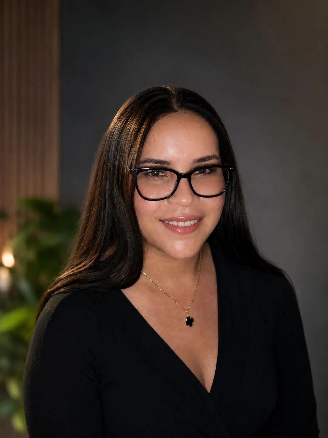 A woman with long dark hair, wearing glasses, a black top, and a gold necklace with a four-leaf clover pendant, smiling at the camera.