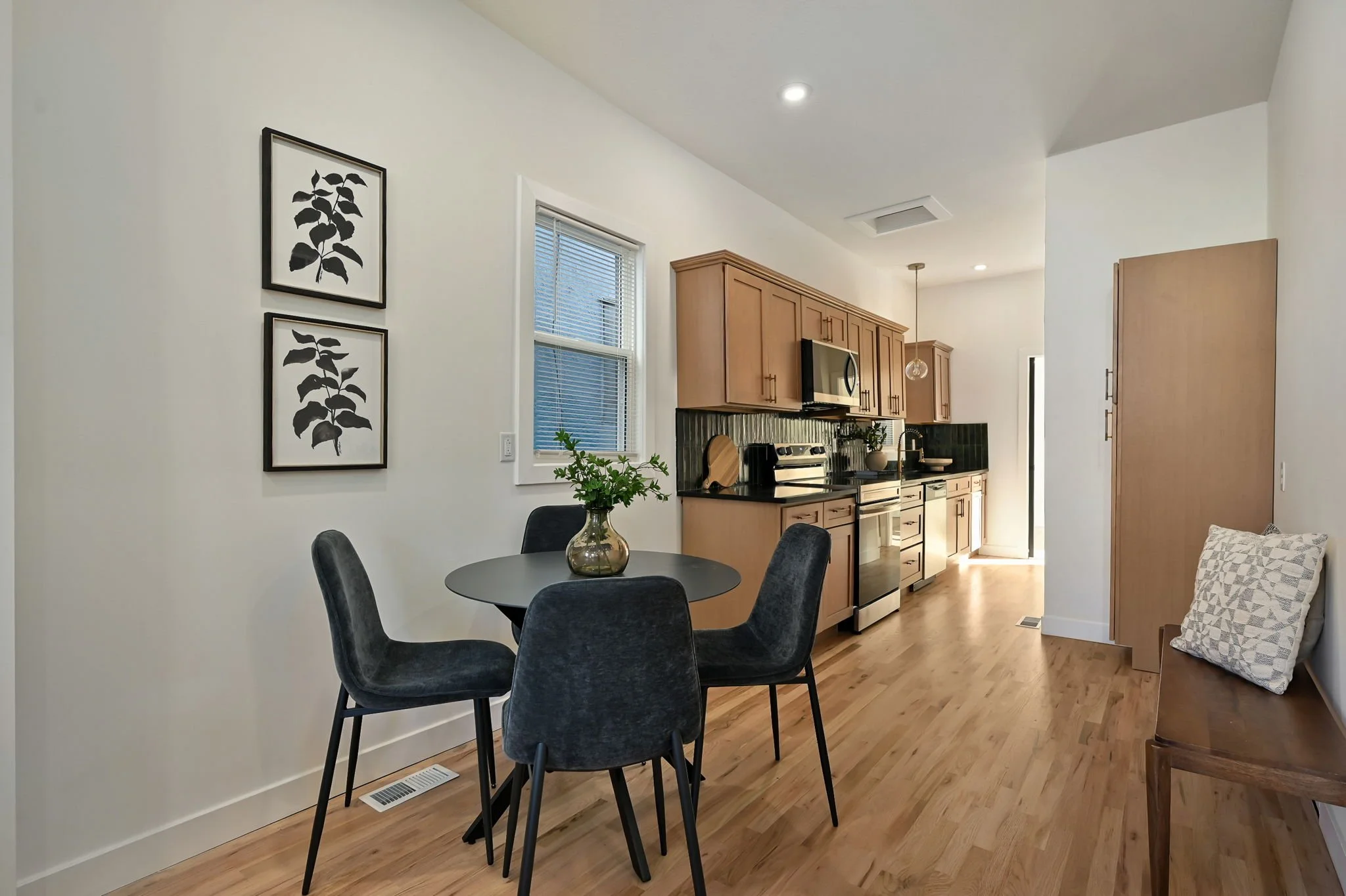 A cozy kitchen and dining area with a black round table surrounded by four dark gray chairs, a kitchen with wooden cabinets, black granite countertops, and stainless steel appliances, a window with blinds, framed black and white leaf art on the wall, and a small corner with a patterned pillow on the floor.