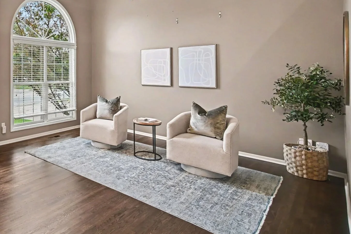 Living room with two beige armchairs, a small round side table with magazines, a large window with white blinds, framed abstract art on the wall, a green potted plant in a woven basket, and a light-colored rug on hardwood floor.