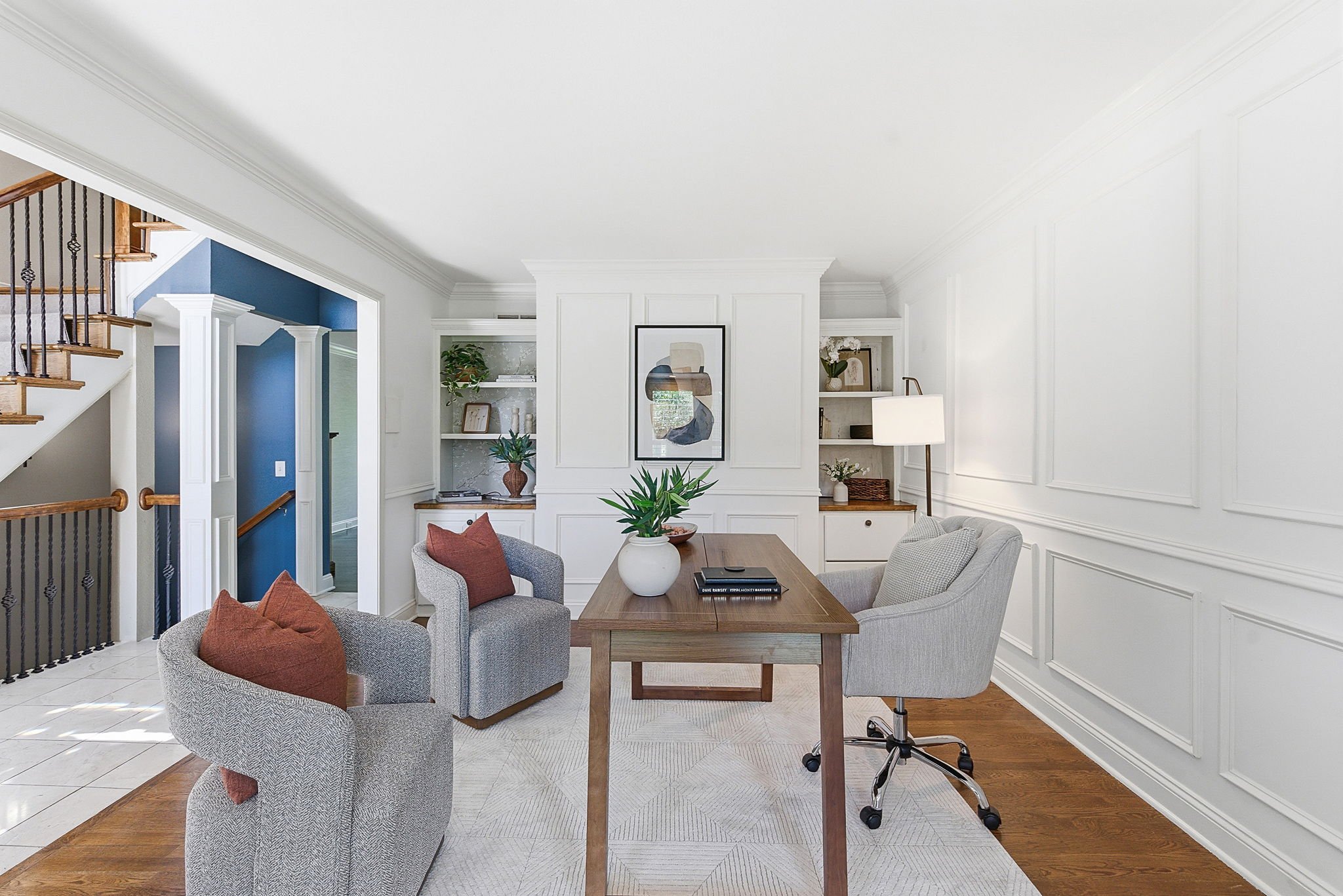 Living room with white walls, wooden floor, and a table surrounded by four grey chairs with pillows. A white vase with green plants and decorative books are on the table. Built-in white shelves with books, plants, and artwork are in the background. There is a large framed abstract art piece on the wall and a white floor lamp.