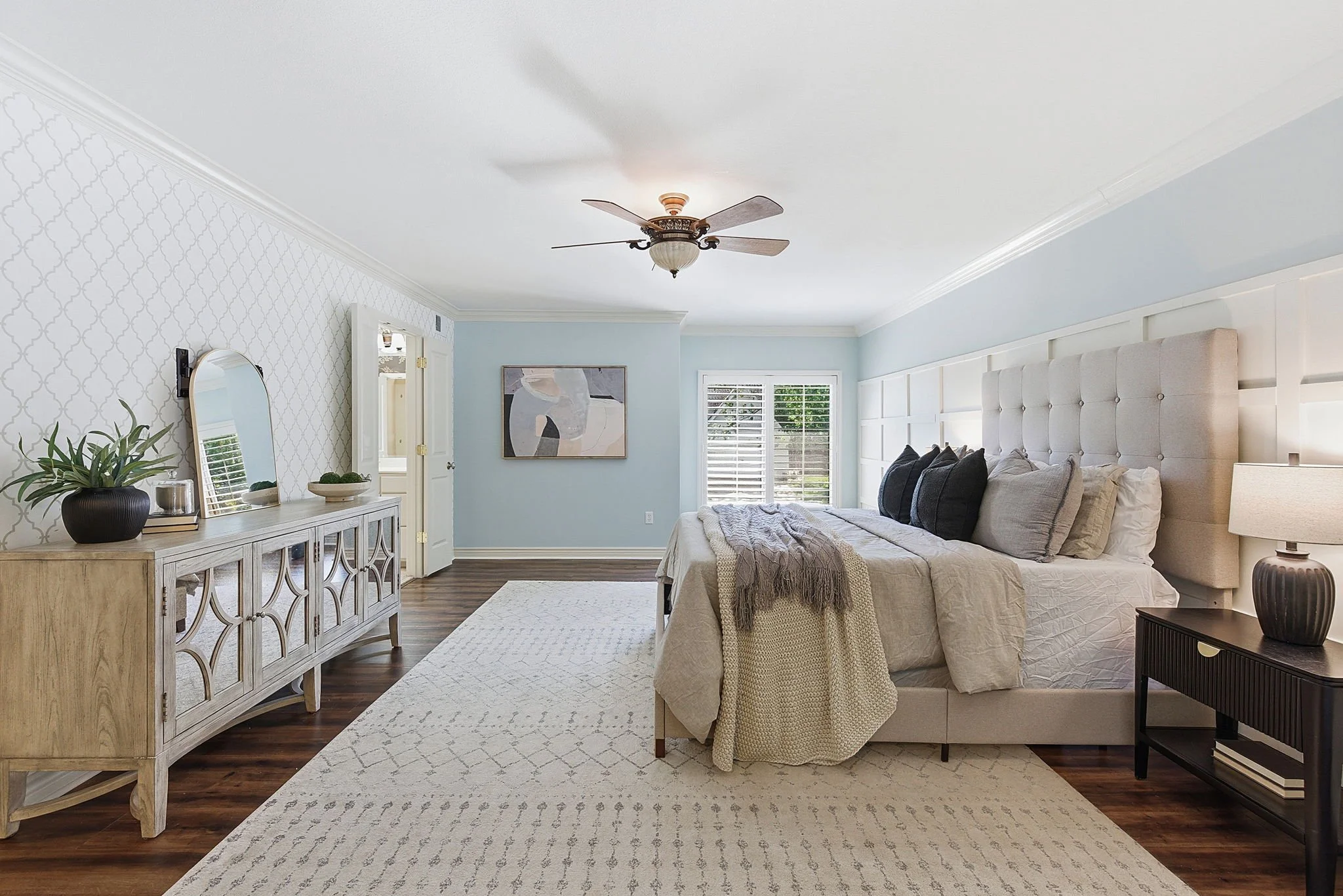 A bedroom with light blue and white walls, a white bed with black accent pillows, a beige headboard, dark wood nightstands, a ceiling fan, a mirror, a white dresser, and a window with white shutters.