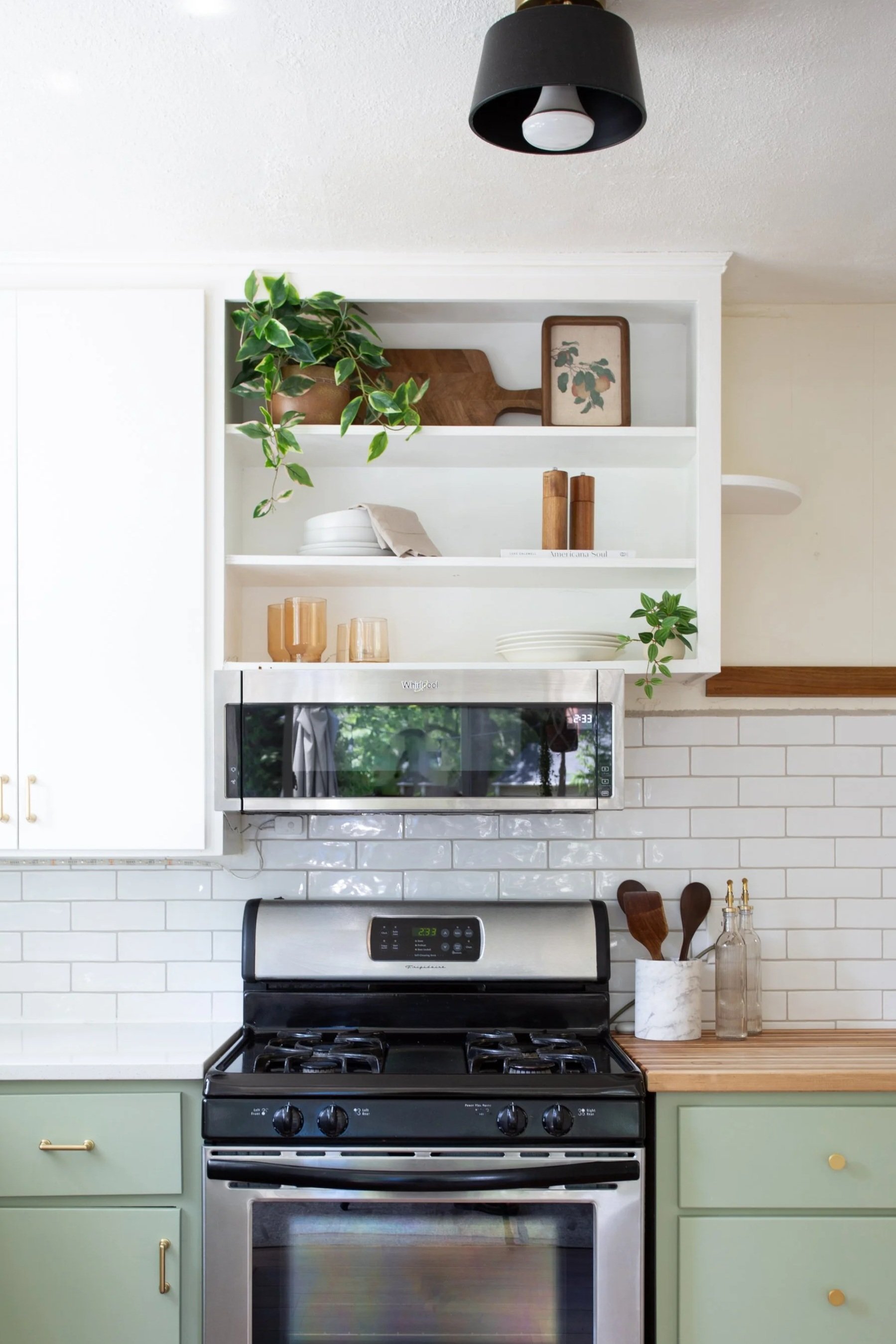 A modern kitchen with a white open shelving above a black stove. The shelves hold a potted plant, wooden cutting boards, ceramic bowls, and glassware. The backsplash is white subway tile, and part of a marble container with kitchen utensils is visible on a wooden countertop to the right of the stove.