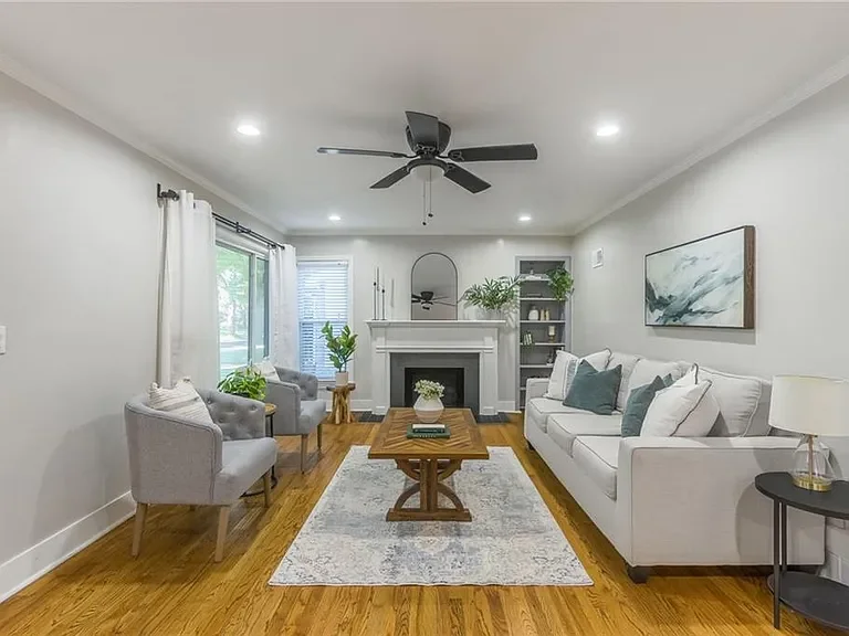 Bright living room with white walls, hardwood floors, a white sofa with throw pillows, two gray armchairs, a wooden coffee table, a fireplace with a mirror above, a window with white curtains, a ceiling fan, and decorative plants.