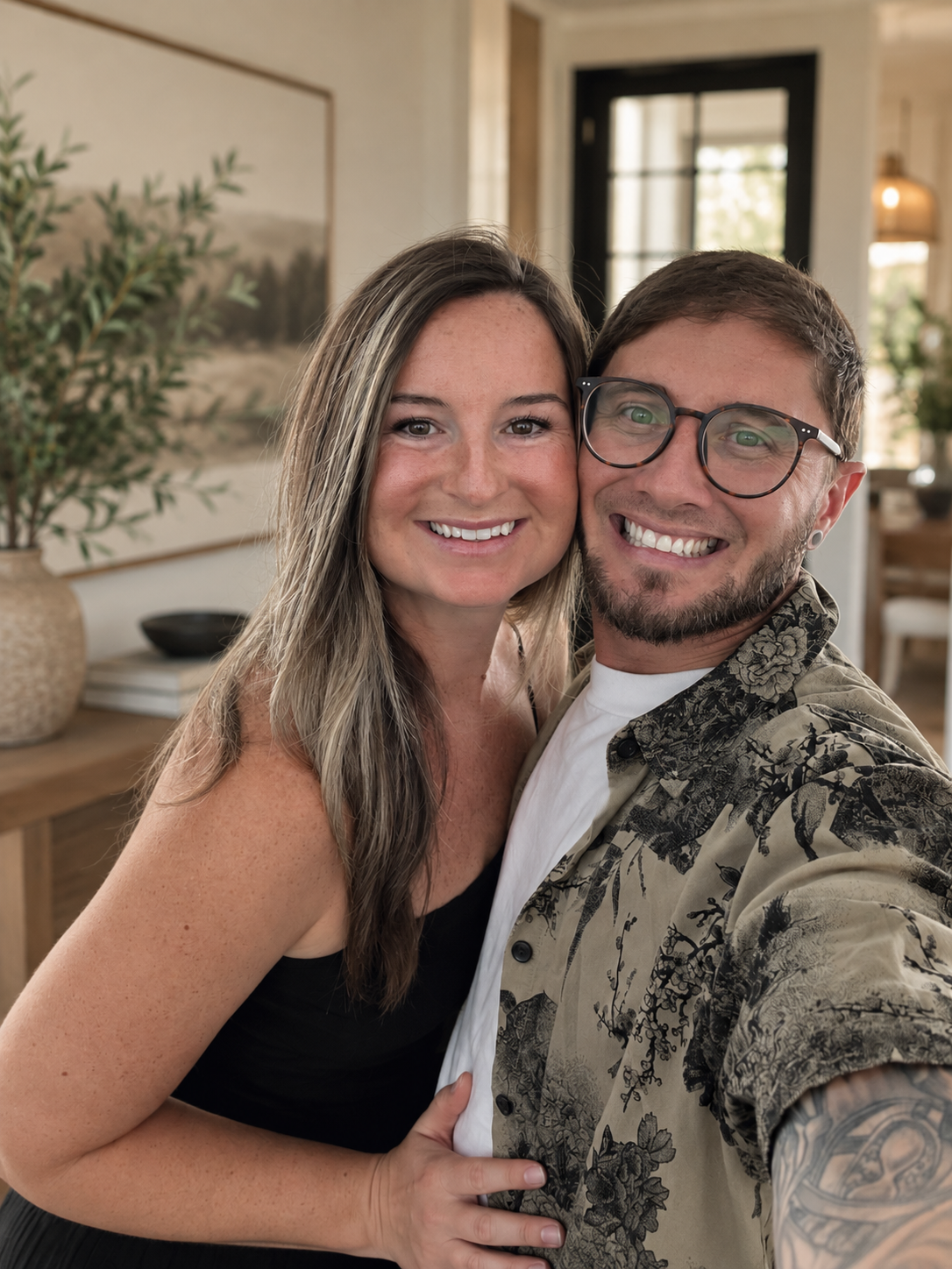 A smiling couple taking a selfie indoors, with the woman on the left having long, wavy hair and the man on the right wearing glasses, a patterned shirt, and a white T-shirt, in a warmly lit room with modern decor.