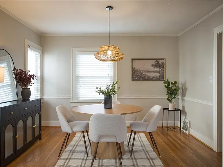 Dining room with a round wooden table, five white upholstered chairs, a black sideboard with a mirror and vase, a hanging pendant light, two windows, a framed landscape picture, and potted plants.