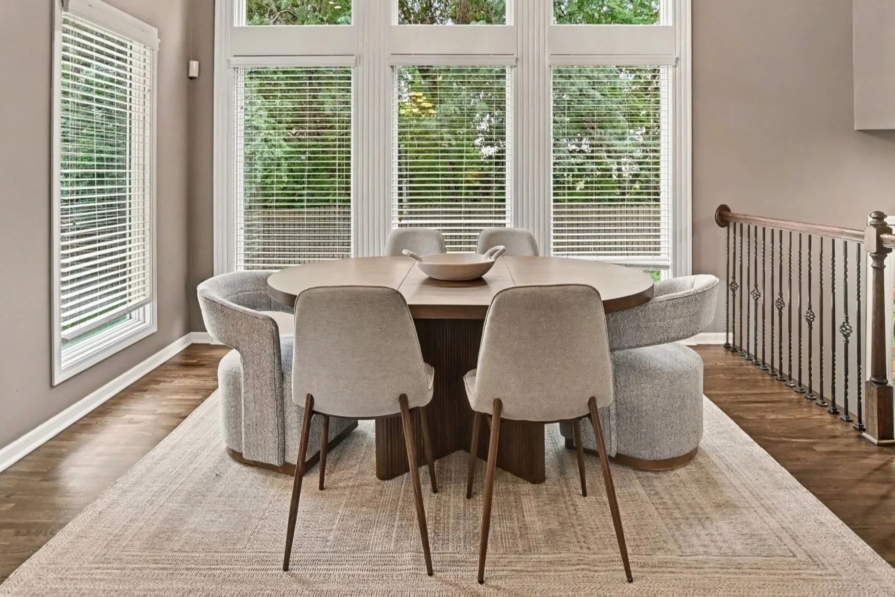 A dining room with a round table surrounded by six cushioned chairs, a large window with white blinds showing greenery outside, wooden flooring, and a beige rug under the table.