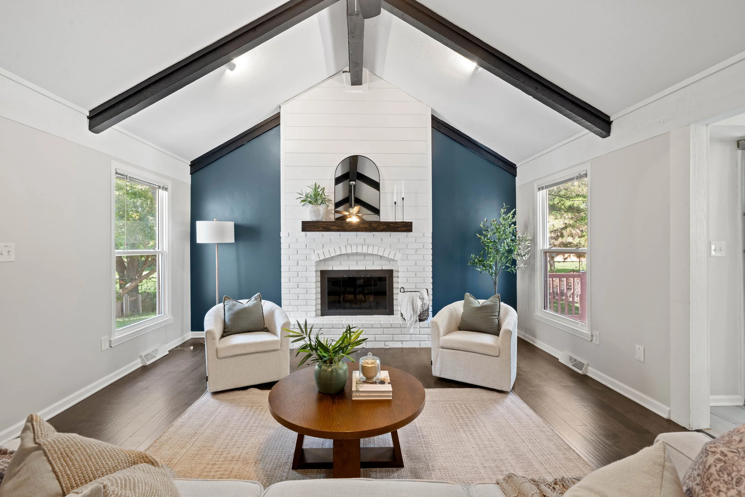 Living room with white walls, blue accent wall behind white brick fireplace, beige armchairs, wooden coffee table with a plant and candles, large windows with trees outside, and decorative pillows.
