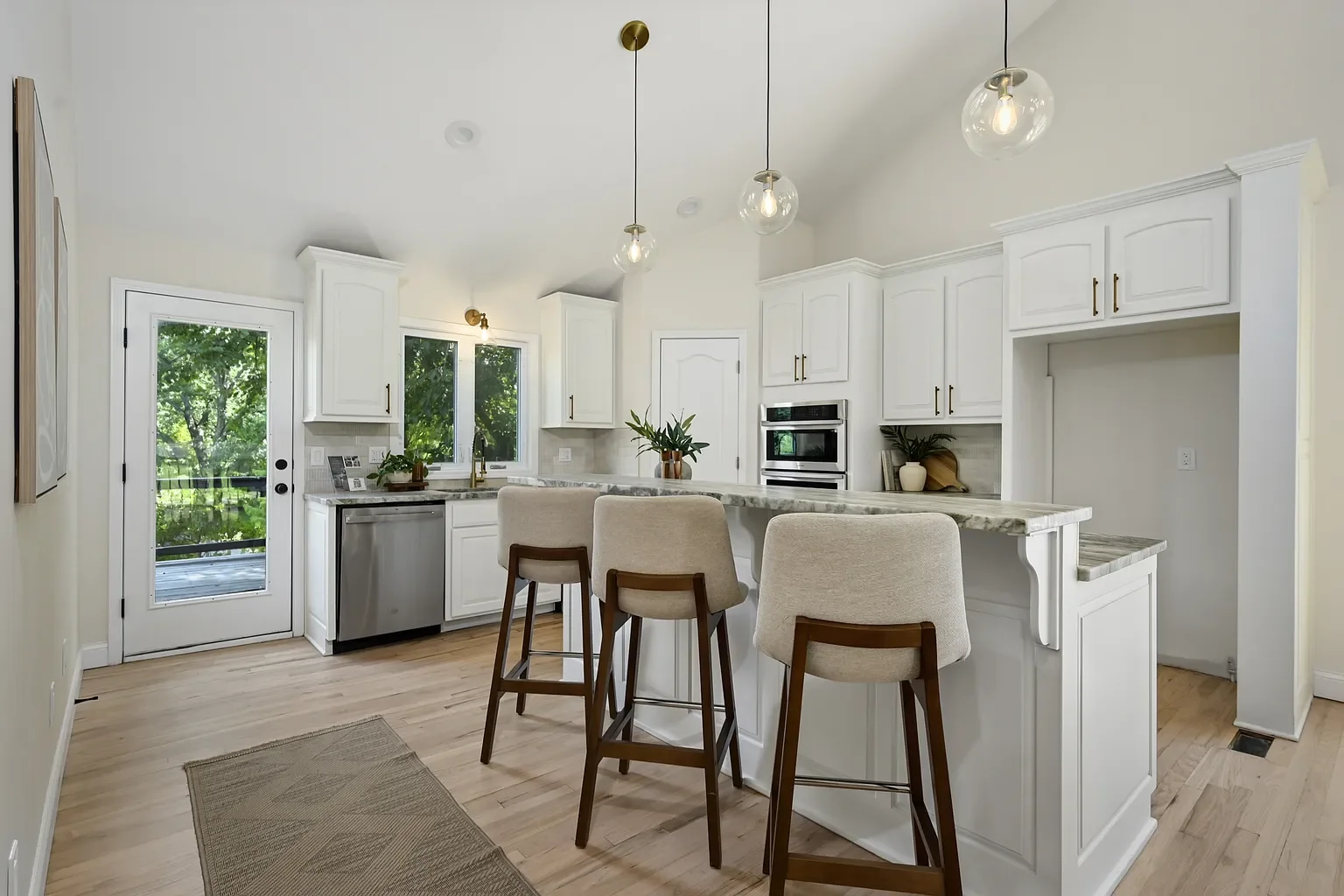 Bright kitchen with white cabinets, marble countertops, a breakfast bar with three beige upholstered stools, and hanging pendant lights, with a door leading to an outdoor deck and large windows showing green trees.