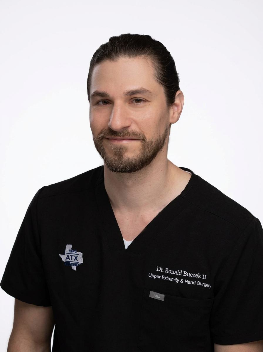 A male doctor with a beard and long hair tied back, wearing black scrubs with embroidered text, standing against a white background.